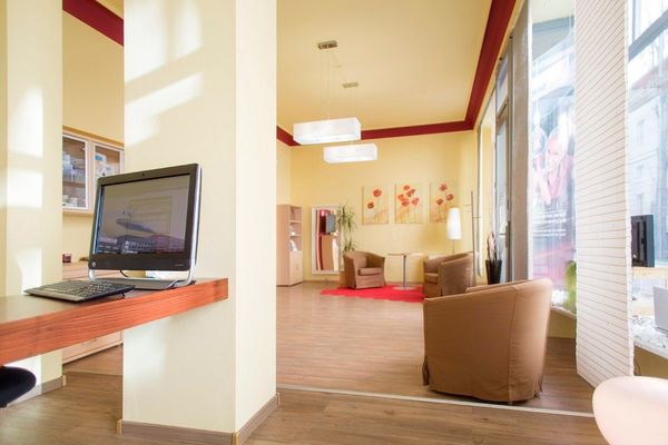 Helles Wartezimmer mit modernen Sesseln, Computerplatz und großer Fensterfront im Tageslicht.