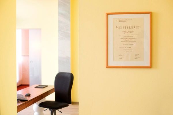 Helles Büro mit Schreibtisch, Bürostuhl und gerahmtem Meisterbrief an gelber Wand.