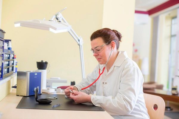 Eine Uhrmacherin prüft eine Armbanduhr mit Stethoskop in einer hellen Werkstatt.